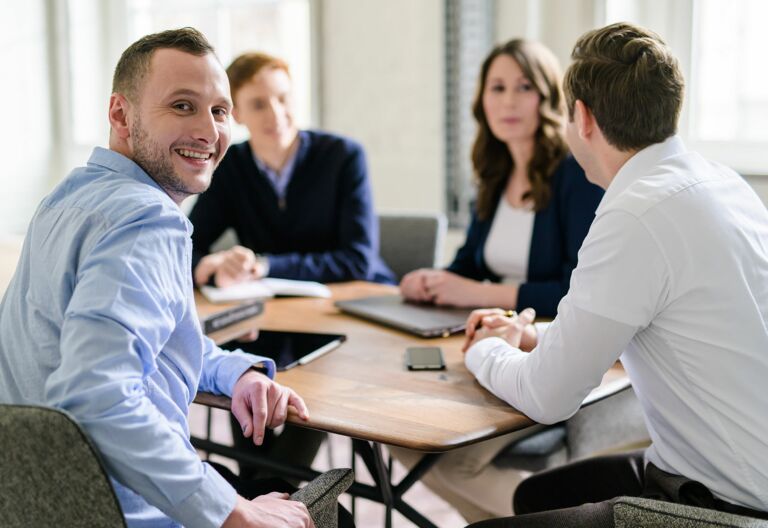 NOVIA GmbH Consulting und Baumanagement - Momentaufnahme Teammeeting an großem Tisch. Junger Kollege schaut über Schulter freundlich in die Kamera.