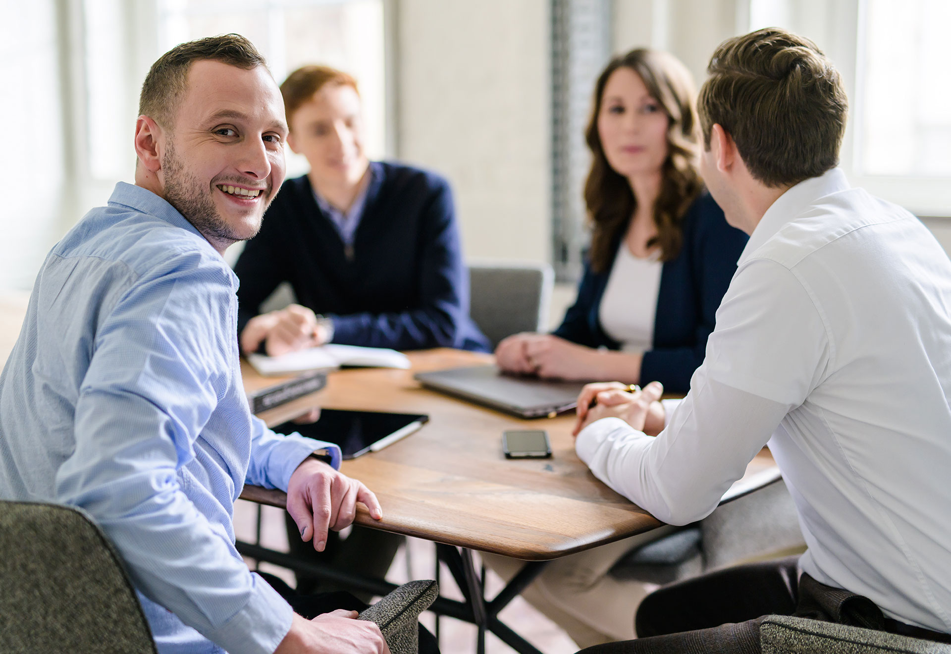 NOVIA GmbH Consulting und Baumanagement - Momentaufnahme Teammeeting an großem Tisch. Junger Kollege schaut über Schulter freundlich in die Kamera.
