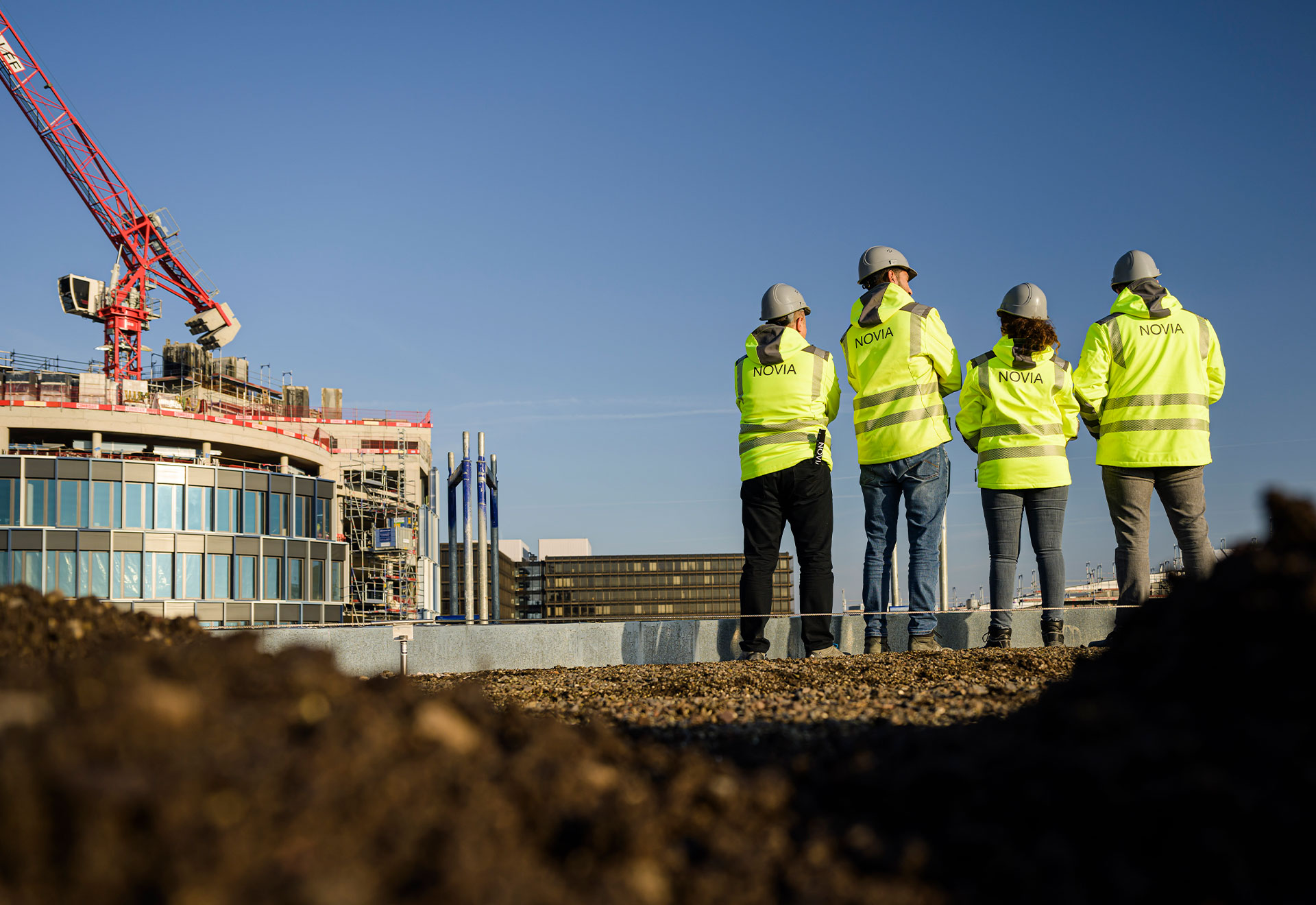 NOVIA GmbH Consulting und Baumanagement - Projektleiter überblicken vom Dach die Baustelle. Sie stehen in gelben Sicherheitsjacken und grauen Bauhelmen vor blauem Himmel mit dem Rücken zur Kamera.